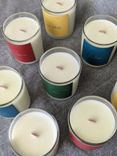 Load image into Gallery viewer, Birds eye view of 7 candles standing up with wooden wicks and colourful labels

