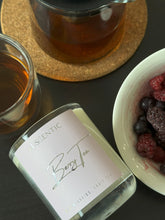 Load image into Gallery viewer, A clear glass candle with a pink label that reads 'LUSCENTIC Berry Tea', placed on a wooden coaster next to a cup of tea and a bowl of berries.