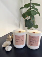 Load image into Gallery viewer, Two white candles with pink labels on a dark surface with a plant in the background.