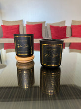 Load image into Gallery viewer, Two black candles with gold text on a reflective surface, with a blurred indoor background.