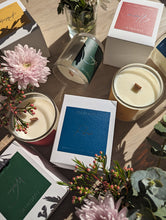 Load image into Gallery viewer, Colourfully labeled boxes in green, blue, red and yellow. Lying flat amongst pink flowers and wooden wick candles in glass jars.