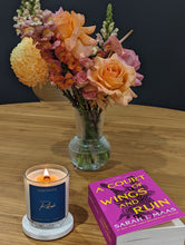 Load image into Gallery viewer, Lit wooden wick candle on the side of a fiction book titled 'A Court of Wings and Ruin' and a bouquet of colourful flowers in a vase