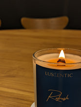 Load image into Gallery viewer, Glass candle with deep, blue label and a lit wooden wick in the middle