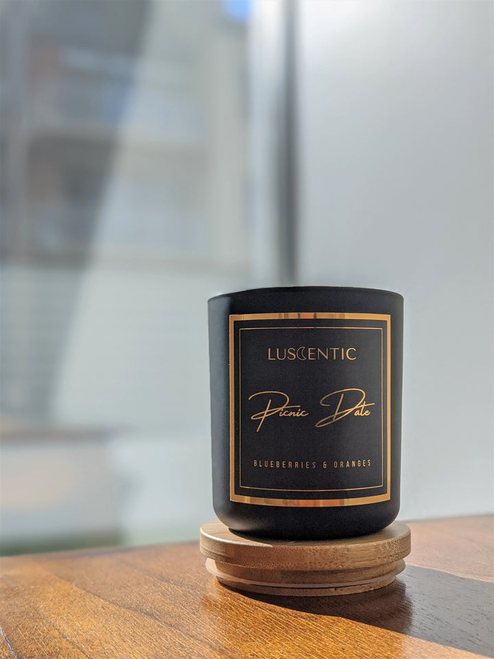 Matte black candle with gold foiling placed on a wooden coaster on a wooden bench. A window is in the background showing a blurred apartment. 